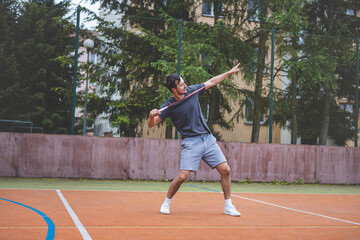 Obraz premium Badminton player celebrates a moment on an outdoor court, holding his racket in an energetic stance. His expression shows excitement and triumph, set against a backdrop of trees and buildings