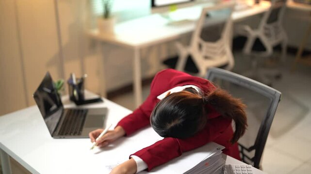 Asian businesswoman in a red blazer slumps over her desk, overwhelmed and exhausted from long hours of work. Papers and a laptop surround her, highlighting the stress of deadlines