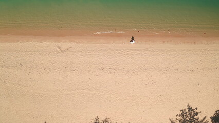 Obraz premium Aerial top down sand beach texture water waves. Woman walking coastline, clear, ideal shore, relax, enjoy outdoor lifestyle travel on summer holiday vacation. Drone view shot. Background