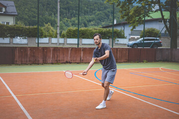 Badminton player focuses on the shuttlecock mid-air during a match on an outdoor court. With his racket raised, he prepares for the next shot, surrounded by an urban and scenic background