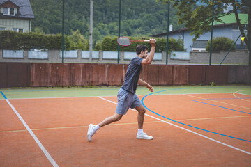 Badminton player focuses on the shuttlecock mid-air during a match on an outdoor court. With his racket raised, he prepares for the next shot, surrounded by an urban and scenic background