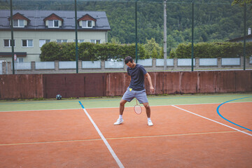 Badminton player returns a shot on an outdoor court, extending his body for a powerful hit. The scene captures athletic focus, with a background of buildings and greenery surrounding the court