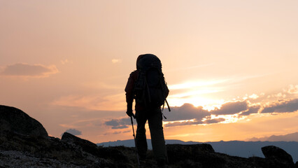 Man hiker porter climber with big backpack and trekking sticks poles raises up to top hilly cliff mountain. Walking at sunrise or sunset sky, sun rays shine. In trekking clothes for hiking. In motion