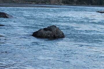 Obraz premium A striking black rock emerges from the clear blue waters of a flowing river, creating a captivating contrast against the vibrant blues. The gentle current surrounds the rock.