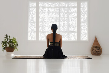 Indian lady, sitting down on a mat in a minimalistic room. She has long braided hair. She is wearing an off shoulder black dress. Facing away. 