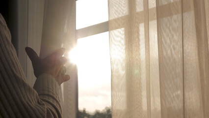 Close up woman hand opening transparent curtain tulle by window to enjoy early morning sunrise at home. Have fun warm energy sun shine sunlight at home and thinking dreaming looking outside at street