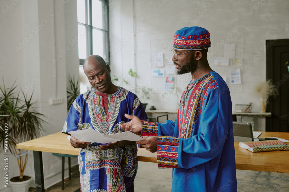 Wall mural African American coworkers in traditional clothes discussing new startup while working offline in modern office