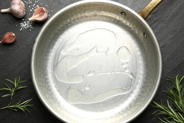 Frying pan with cooking oil, rosemary, garlic and salt on dark textured table, flat lay