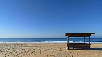 A wooden shelter stands alone on a sandy beach with blue sky and ocean waves in the distance.