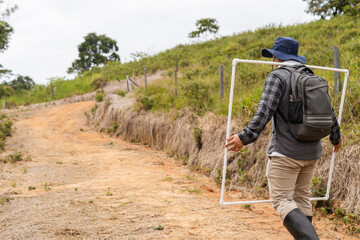 Colombian Latino biologist carrying quadrat frames on his back while walking along a path, preparing to measure pasture growth © Carlostock