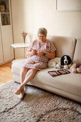 Elderly woman in glasses crocheting at home, a white dog sits beside her