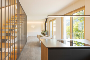 Frontal view of open space with a kitchen island, a table and at the back the sofa and the stove. On the left a modern staircase in wood and metal
