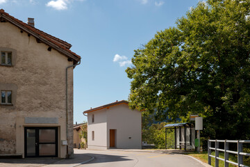 Exterior of the newly renovated house, with the road running past the wall, on a beautiful sunny day with blue skies.  To the left is an old village house and across the street is the bus stop, no one