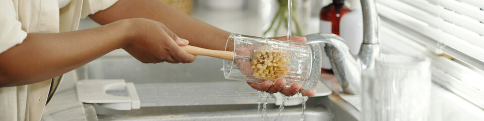 Close-up view of hands washing glass with wooden brush under running water in kitchen sink, showcasing daily chore and cleanliness routine