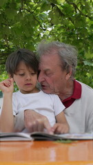 Grandfather and grandson reading a book together outdoors, under the shade of a tree, enjoying quality time and fostering a strong family bond and intergenerational learning