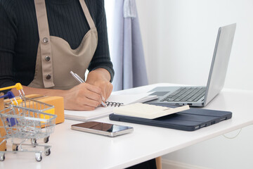 A young businesswoman is multitasking between her laptop and smartphone in her small office. She manages an online store. Monitor orders, inventory, l