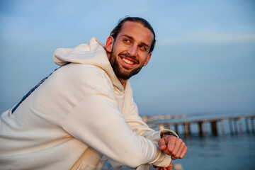 portrait of a handsome and sporty young guy dressed in casual clothes in a seaside location. The model is posing with a smiling expression while standing on a beach at sunset