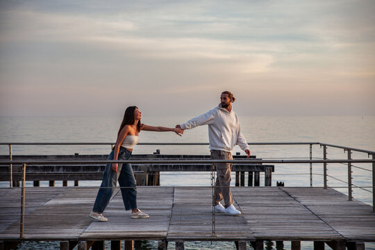 young handsome and charming engaged couple in casual clothes. The two are alone on the pier of a beach at sunset and play with each other holding hands lovingly