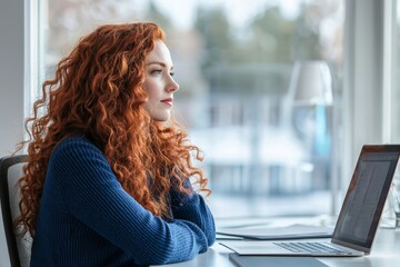 Focused professional woman with curly red hair in modern office setting wearing blue sweater working on laptop and tablet conveying productivity and contentment.