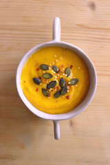 Bowl of creamy pumpkin and potato soup, with pumpkin seeds and chilli flakes toppings. Top view, wooden background.