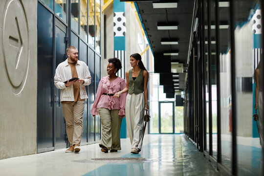 Wide angle shot of diverse group of three colleagues in smart casual clothing walking along business center hallway cheerfully talking after working day, copy space