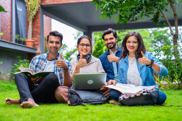 Young Indian college friends sitting comfortably on a green lawn at their college campus, happily sharing smiles as they complete assignments together, surrounded by books and a laptop