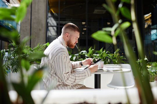 Shot through green living plants of concentrated businessman using digital tablet to work or study remotely sitting at table in cozy sunlit cafe in contemporary office center, copy space