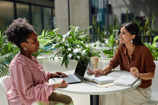 Side view of African American woman discussing work strategy with female colleague while working on laptop at cafe table in contemporary business center, copy space