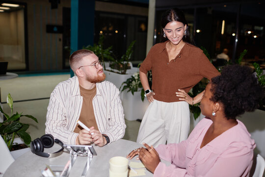 Diverse creative team of two colleagues sitting at cafe table participating in business meeting with supportive female boss checking up on work progress at office center, camera flash