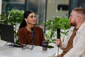 Medium shot of smiling female podcast host engaging in lively discussion with male guest recording broadcast session while sitting at table in cafe with green plants in business center, copy space