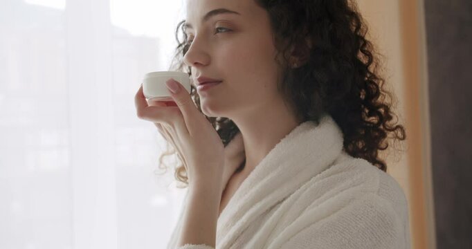 Young woman in a bathrobe holding in her hands and opening a bottle with facial cream and it smells at home. health and wellness concept. Treatment dermatology and cosmetic with moisturiser.