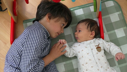 Young boy and baby sibling on a checkered mat, sharing a gaze and a smile, embodying the joy and...