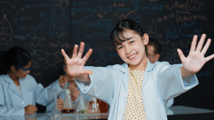 Cute asian scientist waving hand to camera while diverse group doing experiment. Young schoolgirl standing at blackboard with chemical theory written while wearing lab coat at laboratory. Edification.