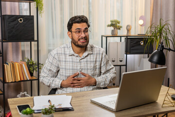 Obraz premium Disappointed Hispanic male freelancer feeling overeating during work process on laptop in home office. Upset Indian businessman freelancer touching stomach and experiencing discomfort and sharp pain.