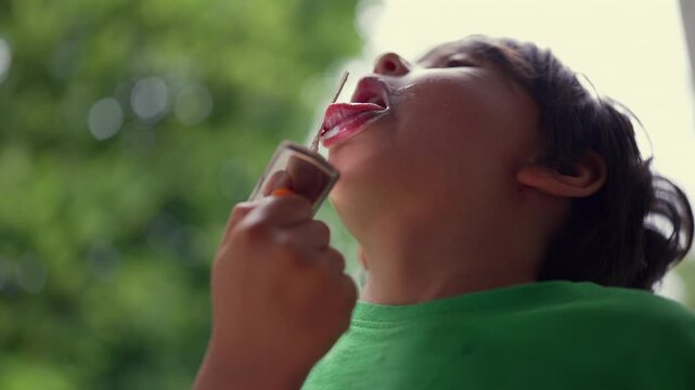 Young boy enjoying ice cream, licking the popsicle in an upward motion. Close-up of a child savoring the dessert outdoors against a green natural background on a warm day