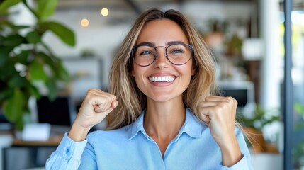 Fototapeta premium A joyful woman with glasses in a light blue shirt is seen clenching her fists in excitement in an office space, capturing triumph and satisfaction.
