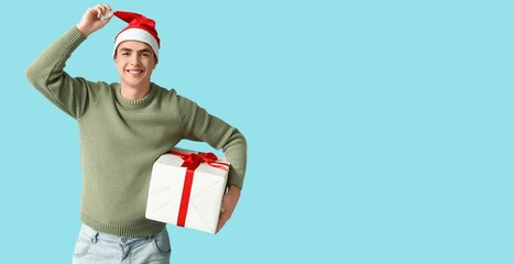 Happy young man in Christmas sweater and Santa hat holding gift box on light blue background with space for text