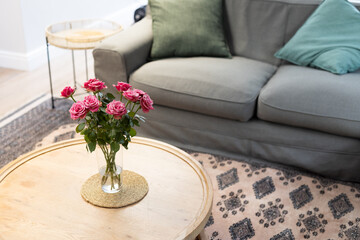 Pink roses in vase on wooden table in cozy living room with sofa, copy space, at home