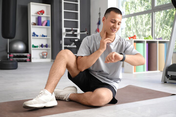 Sporty young man with smartwatch checking pulse on mat in gym