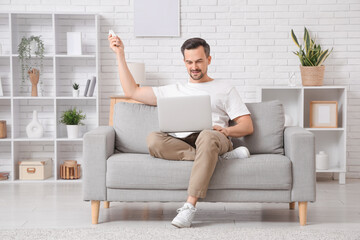 Handsome young man sitting on sofa with air conditioner remote control and using laptop in living...