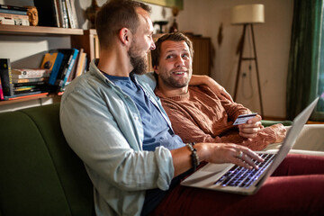 Young male gay couple online shopping with credit card on couch at home
