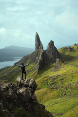 A hiker with his hands up at the top of the mountain views the magnificent landscape with the massive pinnacle of rock. Old Man of Storr, Isle of Skye, Scotland 