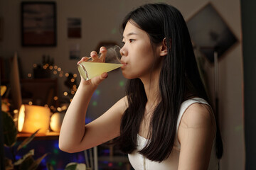 Young Chinese woman with dark long hair sitting in front of camera at home and taking pill dissolved in glass of water
