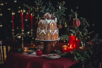 Elegant luxury Christmas table setting for family home party with homemade big gingerbread house, holiday celebration. Red tablecloth, candles, chandelier. Romantic candlelit dinner