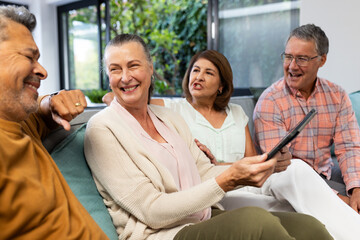 Senior diverse friends sitting on couch, smiling and holding tablet, enjoying conversation, at home