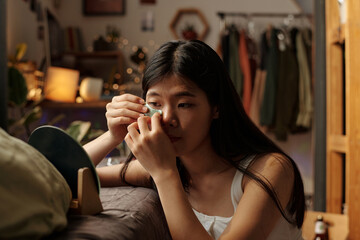 Young woman with baggy skin under eyes putting smoothing patches and looking in mirror while sitting by bed in messy room