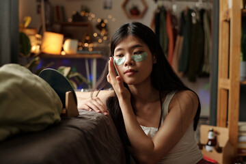 Chinese teenage girl with smoothing patches under eyes looking in mirror while taking care of her...