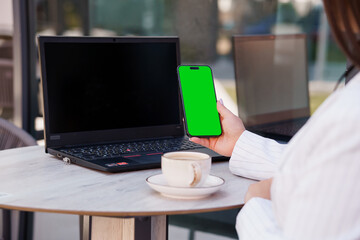 Business woman in a stylish outdoor cafe setting holding a smartphone with a blank green screen while sitting at a table with a laptop and coffee cup
