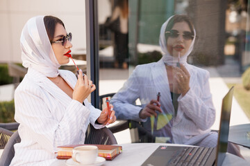 Elegant business woman applying lipstick while working on a laptop with a reflective view