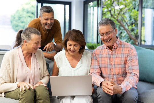 Group of senior diverse friends sitting on couch, smiling and looking at laptop, at home